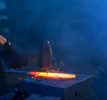 Close up of strong male hands and safety gloves forging molten metal on anvil with hammer. Professional blacksmith working with steel at forge.