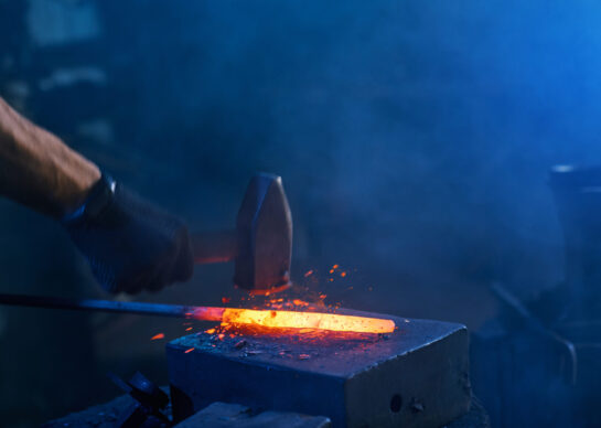 Close up of strong male hands and safety gloves forging molten metal on anvil with hammer. Professional blacksmith working with steel at forge.