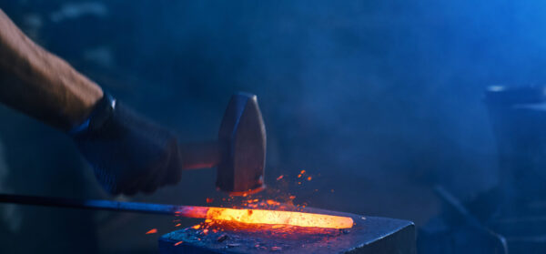 Close up of strong male hands and safety gloves forging molten metal on anvil with hammer. Professional blacksmith working with steel at forge.