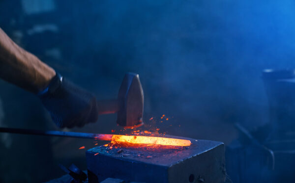 Close up of strong male hands and safety gloves forging molten metal on anvil with hammer. Professional blacksmith working with steel at forge.