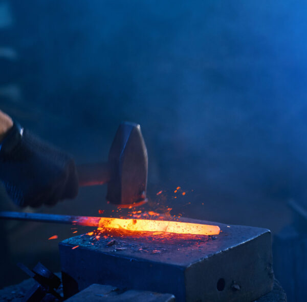 Close up of strong male hands and safety gloves forging molten metal on anvil with hammer. Professional blacksmith working with steel at forge.