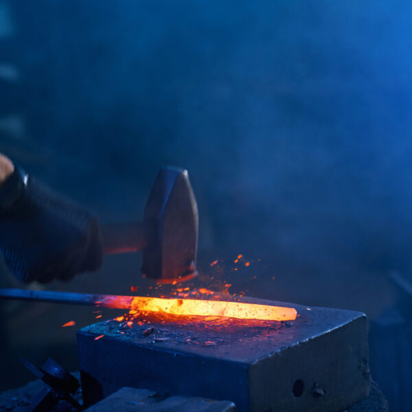 Close up of strong male hands and safety gloves forging molten metal on anvil with hammer. Professional blacksmith working with steel at forge.